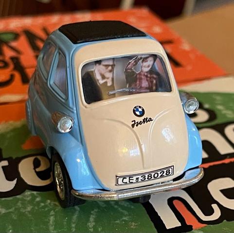 Ein Modellauto, eine hellblau und beige BMW Isetta, das auf einem bunten Buch positioniert ist. Ins Auto wurde Pappfiguren des Ehepaars Schmidt gesetzt. Das Auto steht auf einer Erstausgabe von ”Das steinerne Herz“. Das Autokennzeichen, ursprünglich WI, wurde im Bild bearbeitet. Jetzt CE für Celle.