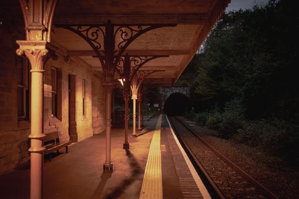 An empty platform at the station just before nightfall; visible are the Victorian canape on the left and in the shadowy section on the right the train tracks lead to the opening of a tunnel in the distance.
