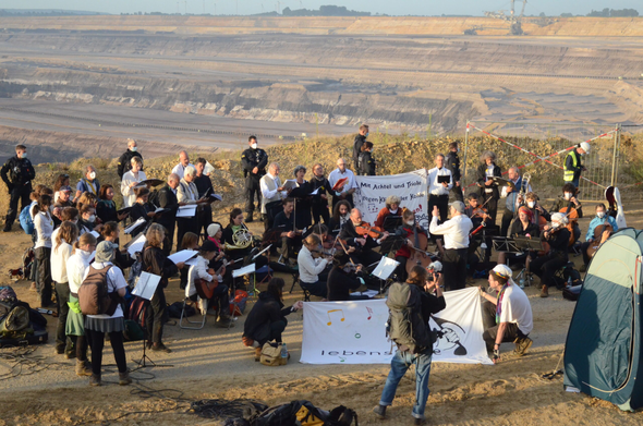 Orchester und Chor, in schwarz-weißer Kleidung, stehen am Rande eines Braunkohletagebaus und musizieren.