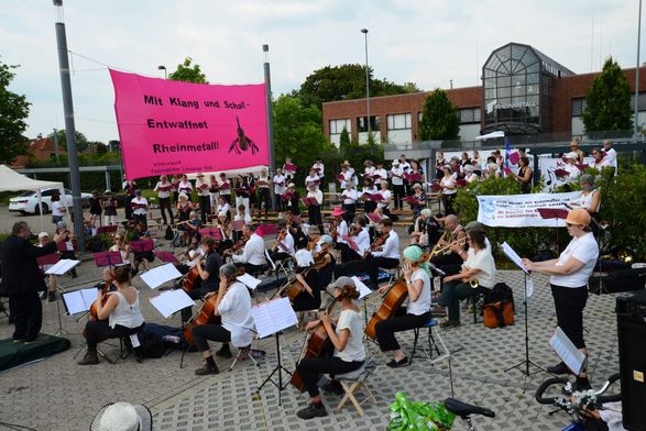 Lebenslaute Musiker*innen vor der Rheinmetall Zentrale.
Über den Musizierenden hängt ein pinkes Transparent mit dem Spruch »Mit Klang und Schall – entwaffnet Rheinmetall«