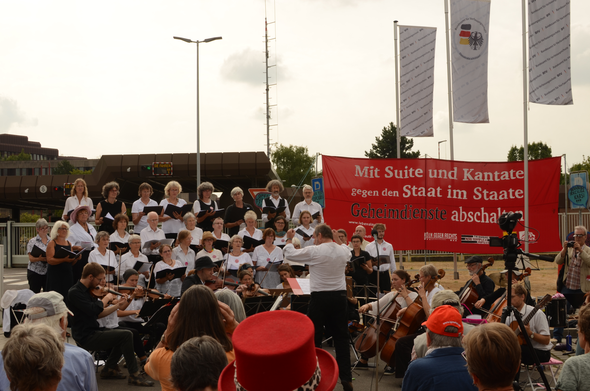Chor und Orchester der Lebenslaute (in schwarz-weißer Konzertkleidung) musizieren vor dem Kölner Verfassungsschutzgebäude.
Daneben ein Transparent: »Mit Suite und Kantate gegen den Staat im Staate. Geheimdienste abschalten«