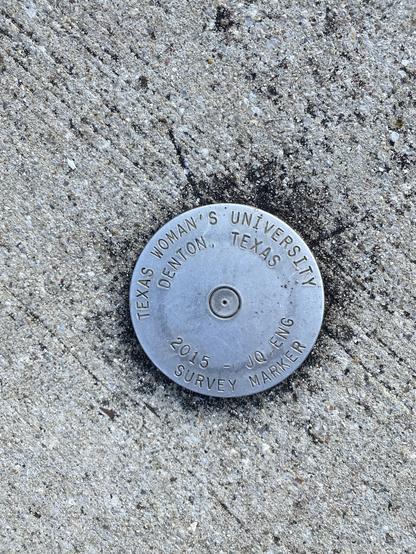 Medallion in concrete. About 3-4 inches wide with “TEXAS WOMAN‘S UNIVERSITY DENTON,  TEXAS 2015 - 10 ENG, SURVEY MARKER”