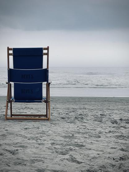 A beach chair in the sand. Beyond, a gray surf under a cloudy sky.