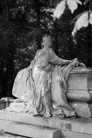 Das schwarzweiss Foto zeigt eine aufwendige Skulptur auf einem Grab, es zeigt eine Frau die sehr grossen Umhang mit einer Hand auf einem Sarg abgelegt hat.