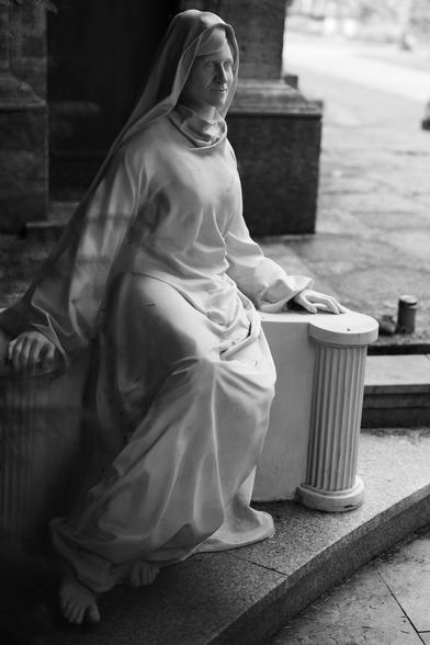 Das schwarzweiss Foto zeigt eine aufwendige Frauenskulptur die auf einem Stein sitzt, sie trägt ein Kopftuch sowie einen Umhang. Die Figur ist sehr fein gearbeitet, besonders die Hände.