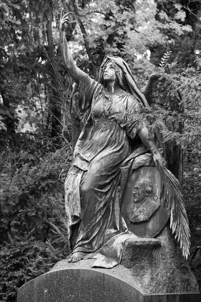 Das schwarzweiss Foto zeigt eine grosse engelartige Skulptur die an einem Stein gelehnt einen Arm nach oben streckt.Sie trägt einen Umhang.