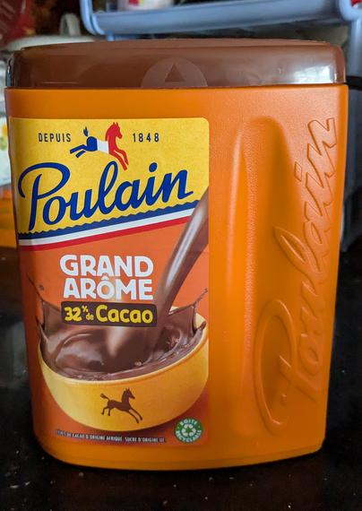 Plastic container of Poulain "grand arôme" hot chocolate mix. The box has "depuis 1848x on top with a horse shaped French flag in between the two words, the word Poulain right under, GRAND ARÔME after that and then 32% Cacao. A bowl filled with a brown liquid can be seen underneath.