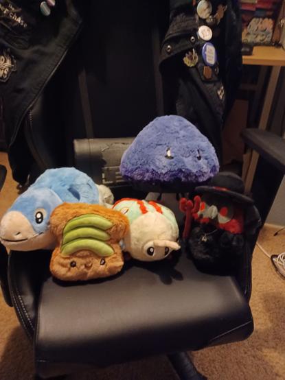 five squishable plushes on a chair: a mosasaurus, an inky cap mushroom, avocado toast, a luna moth, and a demon plague doctor.