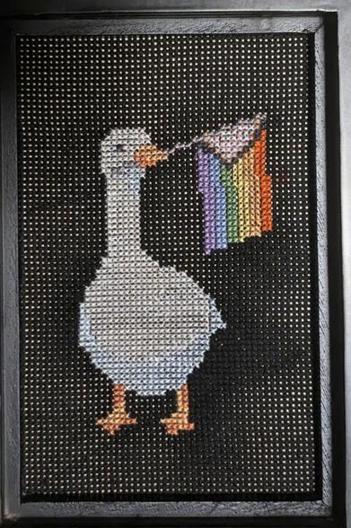 Cross-stitched goose holding a progress pride flag in its beak