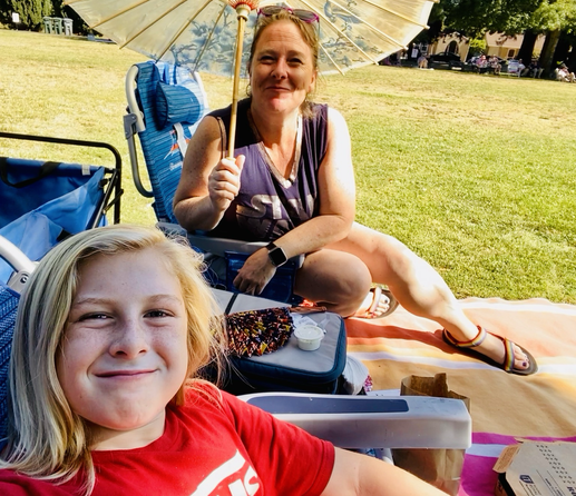 Blond hair boy and mom sitting outside in a park smiling at camera in lawn chairs and with an umbrella.