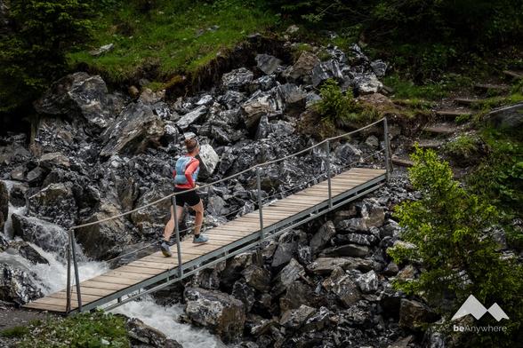 A trail runner in a red shirt and black shorts crosses a narrow wooden bridge over rocky terrain and a small stream, surrounded by lush greenery.