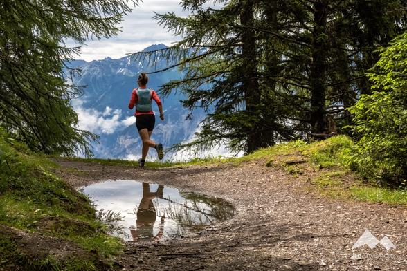 A trail runner in a red shirt and black shorts runs along a forest path, with a mountain view ahead and a reflection in a puddle below, surrounded by tall evergreen trees.