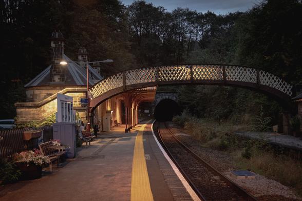 View along the platform at Cromford station. The station buildings are on the left, lit by the streetlighting; an old footbridge crosses the track; in the background is the opening of a tunnel.