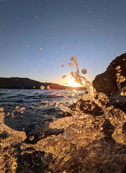 Ocean water splashing - close up, with sunset skies, over mountain ridge & ocean, in the background.