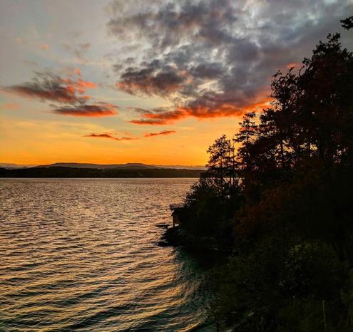 Sunset over the ocean inlet.  Forested cliffs in shadows on the right. In far background are layers of mountain range on the other side of the inlet.