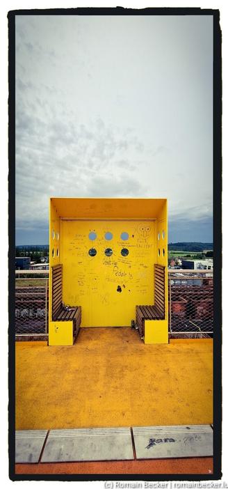 A yellow shelter in front of a dark sky with lots of clouds. Two small benches made out of wood are in the inside.