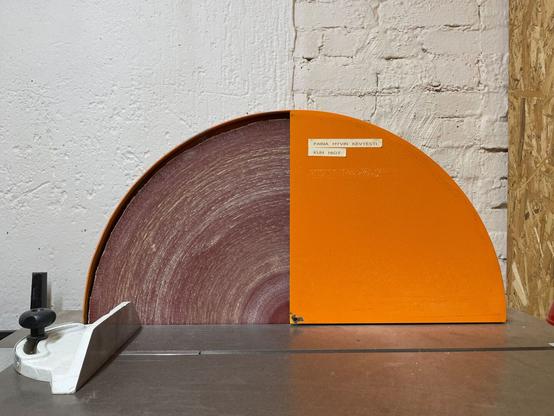 Photo of a sanding machine: a grey table top in the foreground, two quarters of a circle, one reddish, one orange, in the centre of the picture, a white wall behind it, partly painted brick, partly plastered.
