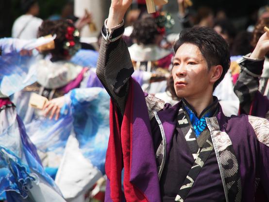 A male dancer strikes a dramatic pose, looking to the side with one arm raised up in front of him. 

The dancer's outfit is multilayered, mainly black, purple and red, with details of iridescent blue, and marbled white and gold.

Behind him are a larger group of dancers out of focus, in white and blue gowns, arms outstretched holding wooden clacker noisemakers.