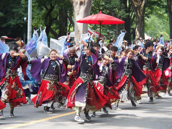 A group of dozens of dancers arrayed in rows along the street, their bodies mostly turned to the right, but twisted, in mid-jump, so they are facing to the left with arms flung up and wide, holding wooden clackers.

The dancer in the previous shot is front and center, the rest of the dancers are a mix of men and women of varying ages, mostly Japanese, but a few likely foreigners as well, including a bald man with dark skin smiling broadly, and most of the dancers are smiling or grinning, some of them hovering slightly above the ground, mid-jump.

Somebody that can't be seen behind the dancers is holding a large red umbrella upright in the middle of the group.

In the background are tall tree trunks and greenery of a roadside park.