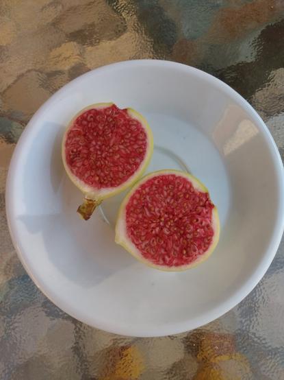 Ein Feige mit saftigem roten Fruchtfleisch in einer gelblich weißen Schale liegt in zwei Hälften geteilt auf einem weißen Teller. 

A fig with juicy red flesh in a yellowish white skin lies on a white plate, divided into two halves.