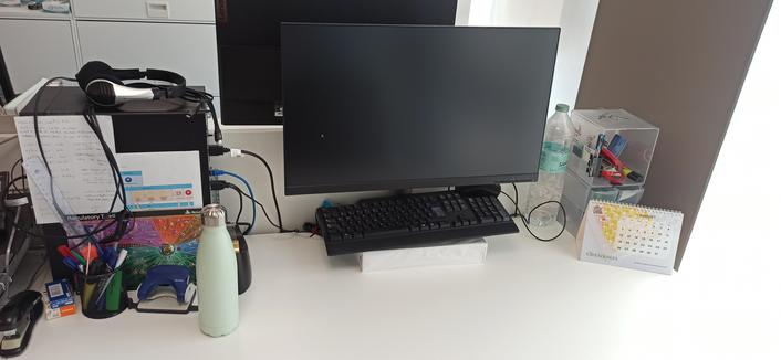 Work desk with a computer on the left, headphones on top of it. A water bottle in front and a screen at the center (above a paper stack). On the right side a calendar and some office objects, staple, marker...