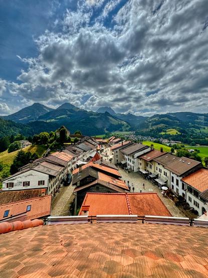 This photo features the picturesque Swiss village of Gruyères, nestled in a valley with red-tiled roofs and cobblestone streets. The village is surrounded by lush green hills and majestic mountains under a partly cloudy sky, creating a serene and idyllic setting.