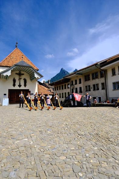 The photo captures a traditional Swiss scene in Gruyères with musicians playing alphorns in a cobblestone square. The square is surrounded by historic buildings, and a small chapel is visible in the background. The backdrop includes a prominent mountain peak, enhancing the cultural and natural beauty of the scene.