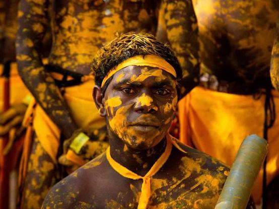 A person covered in yellow and orange paint is looking at the camera, with others similarly painted in the background. They wear headbands and necklaces matching the paint colors.