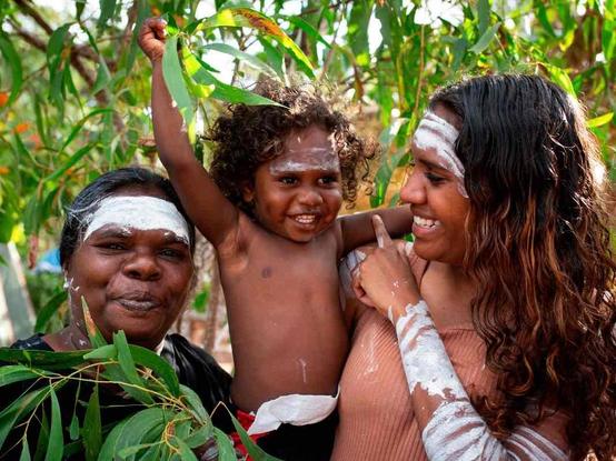 Three people with traditional white paint on their faces and bodies stand outdoors among leafy green branches, smiling and interacting with each other.