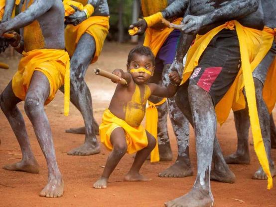 A young child with yellow body paint and a yellow cloth participates in a traditional dance alongside adults with similar body paint and yellow garments.