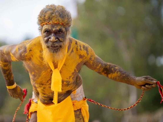 A man covered in yellow body paint, wearing a yellow loincloth and headband, holds red and yellow braided cords while performing in an outdoor setting.