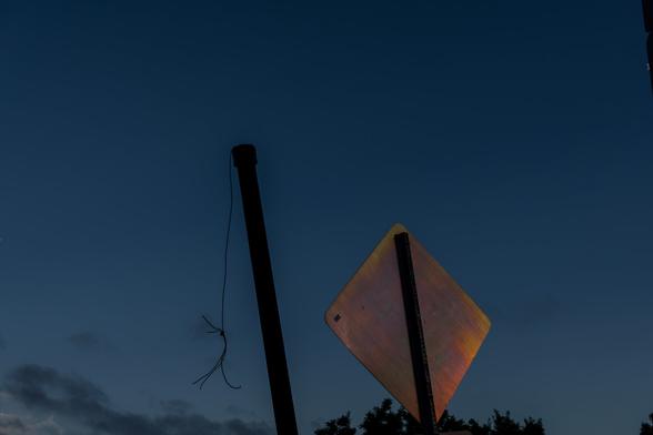 A color photograph taken shortly before sundown. The sun is reflecting off the back of a metal street sign, and a damaged, headless crosswalk pole with protruding wires stands next to it.  A few scattered clouds are visible in the darkening blue sky, and the tops of trees in the park across the street are just visible at the bottom of the frame.