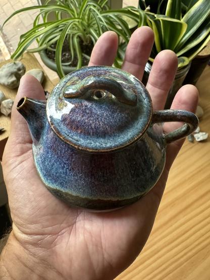 Same teapot with opal glaze in the palm of my hand with house plants in the background.