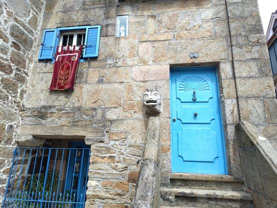 Old stone house in the village, the doors and the window shutters are painted in a bright blue color. 

On the bottom left there's an entrance to the lower floor of the house, with a door and some plants behind a tall blue fence. Over that entrance there's a window with blue shutters, a dark red velvet banner hangs from the window as a decoration for the fair, with an embroidered plant in golden thread. Next to it, there's also a smaller vertical window covered in glass with a figurine inside.

On the right, some big stairs with stone handrails lead to the main door, in the first floor. The left handrail ends in the facade, a smiling animal head made of stone stands over it.