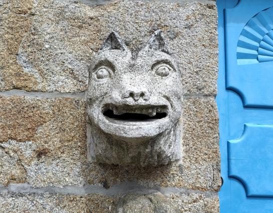 Stone animal head with overall square form, it has small triangular pointy ears, dog-like nose, a big smiling mouth with visible teeth and small funny eyes, apart in the face and looking up.

The inside of the mouth is empty, and connected to the interior of the house, meaning that you can peek outside from the inside of the beast's mouth.