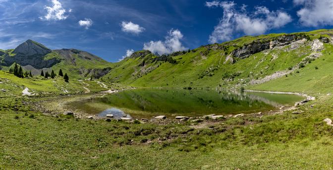 Das Bild zeigt eine malerische Berglandschaft bei klarem Himmel. In der Mitte des Bildes befindet sich ein ruhiger See, der von grünen Hügeln und Bergen umgeben ist. Die Hügel sind mit saftigem Gras und einigen Nadelbäumen bedeckt. Rechts vom See erstreckt sich ein Weg durch die Landschaft. Im Hintergrund ragen steile, felsige Berge empor. Der See spiegelt die umliegenden Hügel und den blauen Himmel wider, was eine friedliche und idyllische Atmosphäre schafft. Die Szene vermittelt ein Gefühl von Ruhe und unberührter Natur.