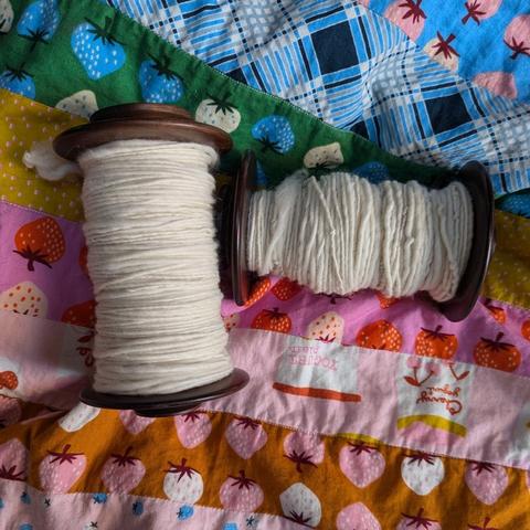 Two singles of white yarn on bobbins sitting at right angles to each other on a colourful quilt. The right one is overspun and noticeably lumpy compared to the left one.