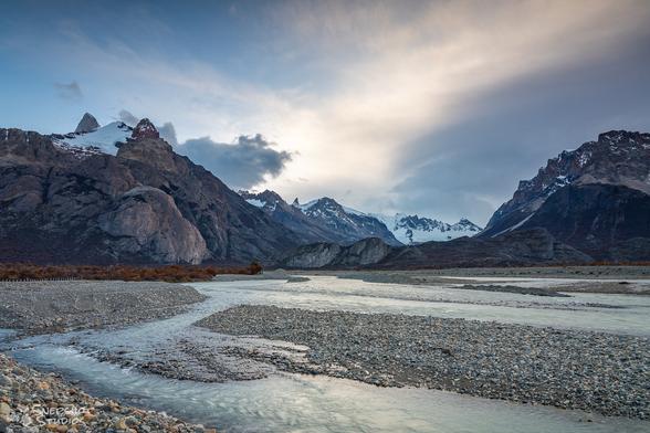Landscape photo of a large gravel plane with multiple branches of a river running through it.  In the back, a high mountain range with some autumn colors.