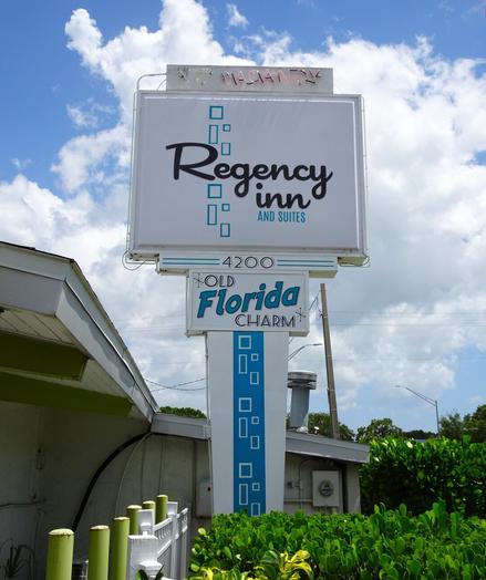 Midcentury modern style sign: Regency Inn and Suites. Old Florida Charm. Turquoise and white sign against a blue sky with puffy white clouds. Neon vacancy light is lit.