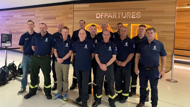A group photo of the first of 67 Aussie firefighters to fly to the US today. They are shown in front of the departure gate at Sydney International Airport wearing their navy polo neck uniform shirts, many in firefighter trousers of navy with neon yellow ankle stripe over black boots.