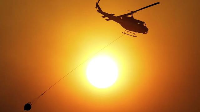 An image of a firefighting helicopter in an orange sky against a hot sun. It is towing a large water bucket to douse the flames of a wildfire.