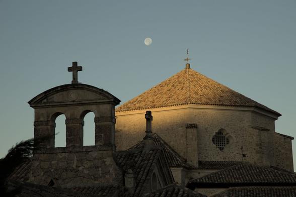 A l'alba, el sol comença a arribar als edificis del casc antic de Cuenca, amb una cúpula i un petit campanar amb una creu. La lluna encara es veu a radera dels dos.