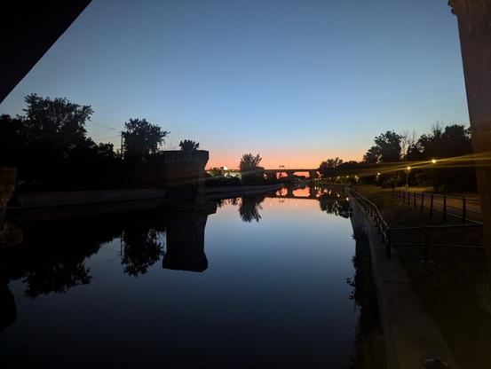 Photo du canal Lachine à l'aube.
Le ciel bleu et rouge à l'horizon se reflète dans l'eau. S'y reflète aussi les lampadaires le long de la piste cyclable ainsi que l'ombre des arbres et d'un pont du loin.