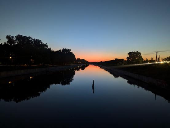 Photo du canal Lachine à l'aube. Une bouée flotte au milieu. Le ciel bleu et rouge à l'horizon se reflète dans l'eau avec l'ombre de arbres en bordure. On y voit les lampadaires de la piste cyclable à gauche, au loin, et des bâtiments industriels à droite.