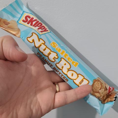 Photograph of a white person's hand holding a wrapped candy bar against a light colored wall. The candy bar has a light blue color on it with the Skippy Peanut Butter logo in the upper left corner. Central to the wrapper, in white text, it reads "Pearson's" with the words "Salted Nut Roll Peanut Butter" beneath it.