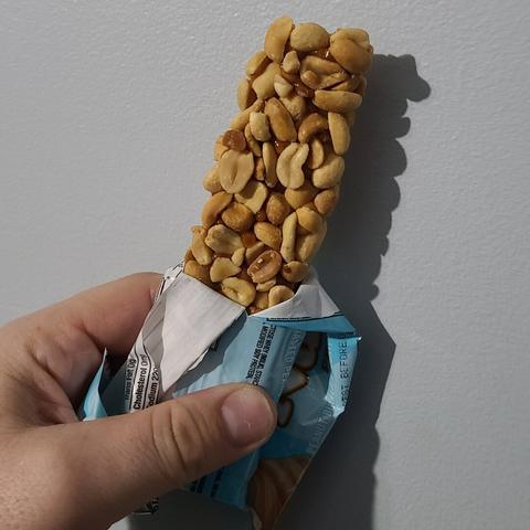 Photograph of a white person's hand holding a partially opened candy bar against a light colored wall. The bar is covered in peanuts with a light brown colored center that's partially visible beneath the peanuts.