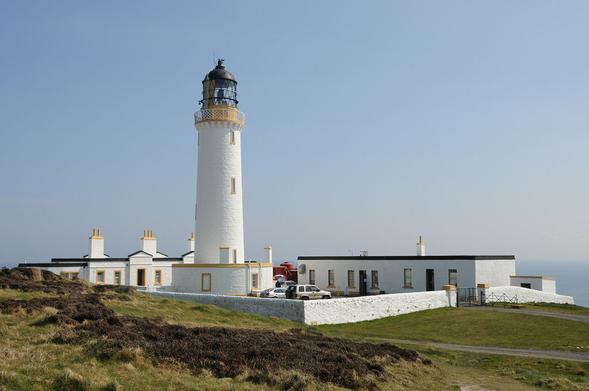 The Mull of Galloway. The image shows a lighthouse just left of the centre of the frame with single-storey white buildings beyond it to the left and right and a low white wall completing a rectangular enclosure this side of it. There is rough ground this side of it and the sea can be glimpsed in the background. The sky is blue.