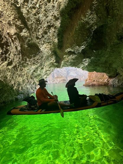 Bob and Christine sitting on a paddle board inside a cave. The bright green water is lit by the reflection of the sunlight into the cave.