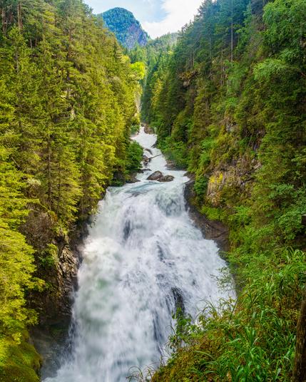 The stunning Reinbach Waterfall tumbles gracefully down rocky precipices, surrounded by vibrant green trees. Gentle mists rise from the cascading water, creating a serene atmosphere as sunlight filters through the forest canopy, illuminating the natural beauty of this Italian gem.