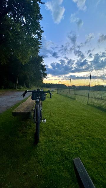 Ein Fahrrad ist gegen eine Holzbank auf einem grasbewachsenen Feld geparkt, mit einem Maschendrahtzaun und Bäumen im Hintergrund bei Sonnenuntergang. Der Himmel ist teilweise bewölkt.